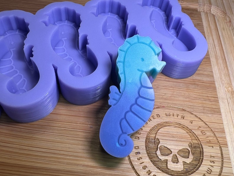 May include: A silicone mold for making seahorse-shaped soap or resin. The mold is purple and has five seahorse cavities. A blue and green seahorse is sitting on top of the mold.