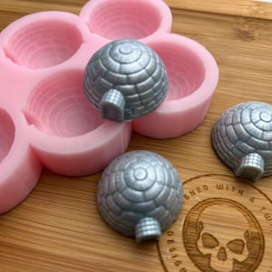 Könnte beinhalten: Eine rosa Silikonform mit sechs runden Vertiefungen, die zum Herstellen von kleinen, kuppelförmigen Süßwaren oder Seifen gedacht ist. Drei silberfarbene, kuppelförmige Süßwaren sind außerhalb der Form zu sehen.