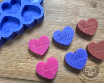 Op de afbeelding: Zes hartvormige zeepjes met de woorden "Thankyou" erop. De zeepjes zijn in tinten paars, roze en bruin. De zeepjes liggen op een houten oppervlak.