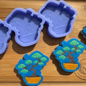 May include: Two purple silicone molds for making resin crafts, each with a design of a bonsai tree in a pot. Two finished resin crafts are also shown, one with a blue background and the other with a green background.
