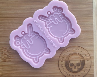 Könnte beinhalten: Eine rosa Silikonform mit zwei Abdrücken eines Comic-Charakters mit erhobenen Armen. Die Form ist mit "Designed with a Twist" beschriftet und hat ein Totenkopf-Logo mit demselben Text.