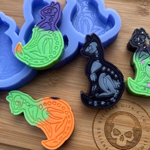 May include: A silicone mold with four cavities shaped like skeletons. The mold is purple and blue. There are four finished resin pieces in the colors green, black, and orange. The resin pieces are sitting on a wooden surface. The text "Designed with a twist" is visible on the surface.