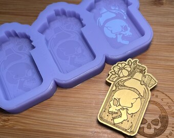 Pode incluir: Um molde de silicone roxo com três cavidades em forma de garrafas com crânios dentro. Uma garrafa de madeira com um crânio dentro e detalhes florais é mostrada ao lado do molde.