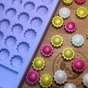 Puede incluir: Un molde de silicona morado con 36 cavidades en forma de estrella, junto a una superficie de madera con 15 fundidos de cera en forma de estrella en rosa, blanco y amarillo.