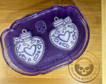 Könnte beinhalten: Lilafarbene Harzschale mit zwei herzförmigen Trankflaschen-Designs und einem Totenkopf. Auf der Schale steht der Text "DESIGNED WITH A TWIST". Ein Holzuntersetzer mit einem Totenkopf-Design und demselben Text ist ebenfalls vorhanden.