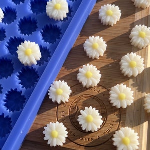 May include: White daisy-shaped embellishments with yellow centers are scattered on a wooden surface. A blue silicone mold with daisy-shaped cavities is visible. The daisies are likely for crafts or decorations.