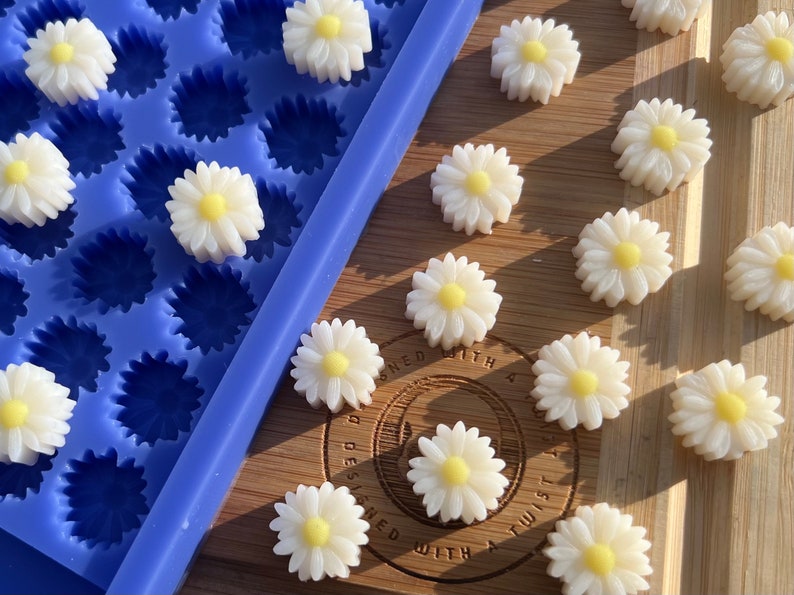 May include: White daisy-shaped embellishments with yellow centers are scattered on a wooden surface. A blue silicone mold with daisy-shaped cavities is visible. The daisies are likely for crafts or decorations.