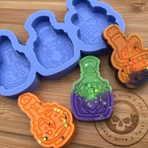 May include: A silicone mold for making potion bottles, with four different designs. The mold is purple and the bottles are green, orange, and purple. The bottles are filled with glitter and have a spider design. The mold is on a wooden surface with a skull logo that says "Designed with a Twist".