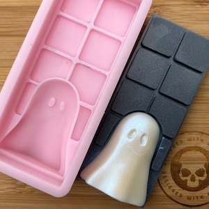 Puede incluir: Un molde de silicona rosa con forma de fantasma y una barra de jabón con forma de fantasma en blanco y negro. La barra de jabón está hecha en el molde y tiene un acabado brillante. El molde tiene una cuadrícula de cuadrados. La barra de jabón tiene un logotipo de calavera con el texto "Designed with a Twist".