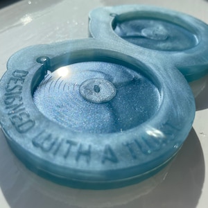 May include: Two light blue coasters with a glittery finish. The coasters are designed with a raised rim and the words "Designed with a Tilt" are inscribed on the side.