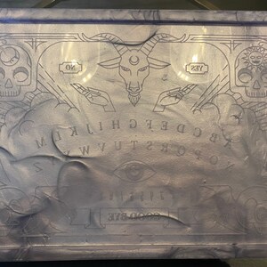 Pode incluir: Um tabuleiro Ouija retangular, cor lavanda, com acabamento brilhante. O tabuleiro apresenta letras, n&uacute;meros e s&iacute;mbolos gravados, incluindo uma cabe&ccedil;a de bode, caveiras e as palavras "SIM" e "N&Atilde;O". O design &eacute; emoldurado por bordas decorativas.