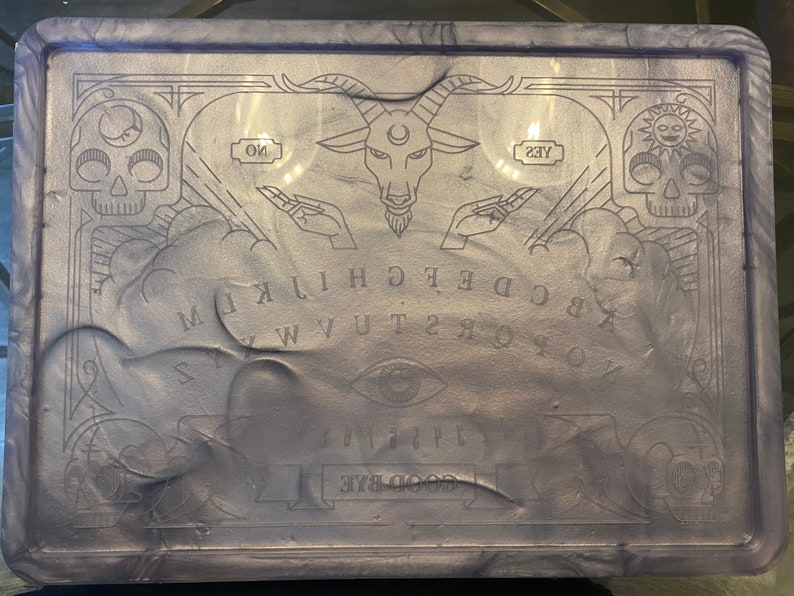 Pode incluir: Um tabuleiro Ouija retangular, cor lavanda, com acabamento brilhante. O tabuleiro apresenta letras, n&uacute;meros e s&iacute;mbolos gravados, incluindo uma cabe&ccedil;a de bode, caveiras e as palavras "SIM" e "N&Atilde;O". O design &eacute; emoldurado por bordas decorativas.