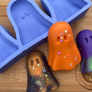 May include: A silicone mold with four ghost shapes, and three finished ghost-shaped candles. The candles are orange with white sprinkles, purple with green and yellow accents, and black with gold glitter and white stars.