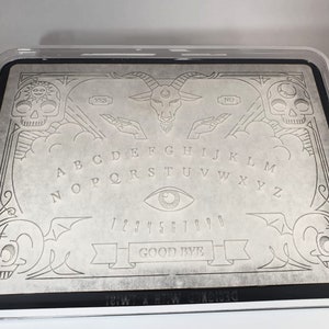 May include: A grey Ouija board with a black border, featuring engraved letters, numbers, and symbols. The design includes skulls, a goat head, and the words "YES," "NO," and "GOOD BYE." The board is in a clear plastic case.