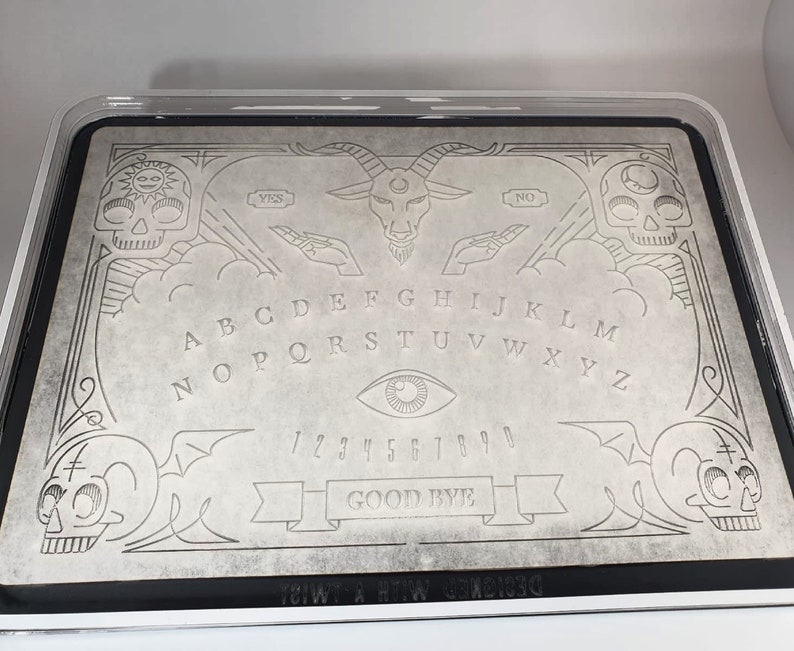 Pode incluir: Um tabuleiro Ouija cinza com uma borda preta, com letras, n&uacute;meros e s&iacute;mbolos gravados. O design inclui caveiras, uma cabe&ccedil;a de bode e as palavras "YES", "NO" e "GOOD BYE". O tabuleiro est&aacute; em uma caixa de pl&aacute;stico transparente.