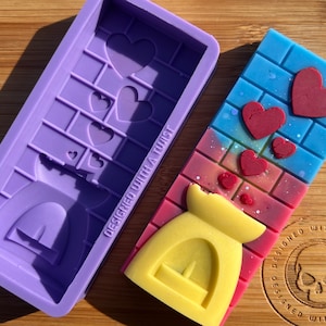 Op de afbeelding: Paarse siliconen mal om een kaars in de vorm van een open haard met harten te maken. De mal ligt naast een afgewerkte kaars met een gele was open haard en rode harten op een blauw-roze bakstenen achtergrond. De kaars heeft de tekst "Designed with a Twist" aan de zijkant.