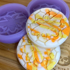 Könnte beinhalten: Drei runde, weiße Seifenstücke mit einem Wabenmuster und einem Bienenbild. Die Seifenstücke sind gelb, orange und weiß mit goldenem Glitzer. Die Seifenstücke befinden sich in Silikonformen mit einem Bienendesign.