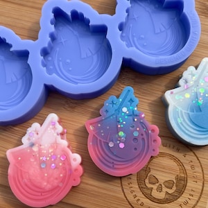 Puede incluir: Un molde de silicona con tres diseños de unicornio. El molde es morado y los diseños están delineados en blanco. Hay tres piezas de resina de unicornio terminadas, una rosa, una azul y una morada, con purpurina incrustada en la resina.