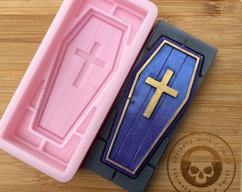 Könnte beinhalten: Rosa Silikonform in Form eines Sarges mit einem Kreuz auf dem Deckel. Neben der Form ist ein lila-goldener Sarg mit einem Kreuz zu sehen. Die Form ist mit einem Twist entworfen.
