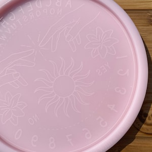 May include: Pink circular silicone mould with a sun, two hands, and two flowers etched into the surface. The mould also has the letters of the alphabet and numbers 0-9 around the edge. The mould is on a wooden surface.