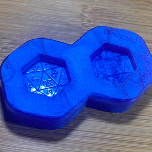 Könnte beinhalten: Blaue Silikonform zum Herstellen von zwei Würfeln. Die Form hat auf jeder Seite ein erhabenes Design eines 20-seitigen Würfels. Die Form ist mit "Designed by Wicked Dice" beschriftet.