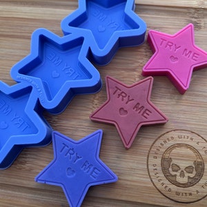Peut inclure: Cinq moules en silicone en forme d'étoile, trois sont bleus et deux sont violets. Les moules ont les mots "Try Me" et un motif de cœur à l'intérieur. Les moules sont sur une surface en bois avec un logo de crâne qui dit "Designed with a Twist".