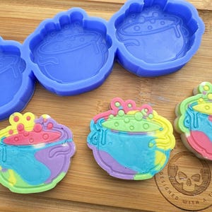 May include: A set of four silicone moulds for making cauldron shaped soaps. The moulds are blue and feature a cauldron with bubbles. There are two finished soaps in the image, one is green and blue and the other is pink and yellow.