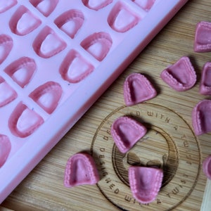 May include: Pink silicone mold with 36 individual cavities shaped like teeth.  The mold is on a wooden surface with several pink gummy teeth scattered around it.  The text "Designed with a Twist" is visible on the wooden surface.