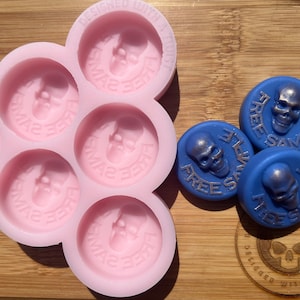 May include: Pink silicone mold with six round cavities, each featuring a skull design with the text "Free Sample".  Three blue round wax melts with a skull design and the text "Free Sample" are on a wooden surface.