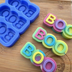 May include: A blue silicone mold with six cavities shaped like the letters "BOO" and three finished colorful soap bars that spell "BOO" in a rainbow swirl pattern.