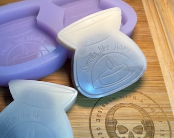 Op de afbeelding: Twee transparante siliconen mallen voor het maken van kaarsen in de vorm van een kleine keramische pot. De mallen liggen op een houten oppervlak. De potvorm heeft de tekst "Smells like Home" en een kleine kaarsvlam afbeelding.