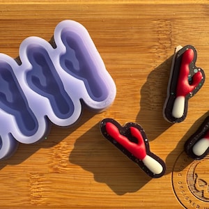Puede incluir: Un molde de silicona morado con cuatro formas de cactus. Se muestran tres piezas de resina en forma de cactus rojo y blanco junto al molde.