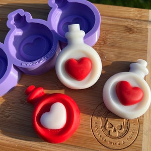 May include: Three white and red heart-shaped potion bottles with a purple silicone mold in the background. The mold has the text "Designed with a Twist" on it.