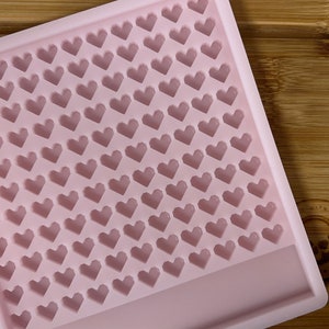 Op de afbeelding: Roze siliconen mal met een raster van kleine hartvormige holtes. De mal heeft een rechthoekige vorm en een glad oppervlak.