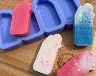 Op de afbeelding: Vier siliconen mallen om zeep of was smelt te maken in de vorm van een fles met het woord "FRESH" op het etiket. Twee van de mallen zijn gevuld met roze, witte en blauwe zeep of was smelt. De mallen liggen op een houten oppervlak.