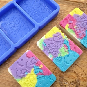 May include: A silicone mold with three rectangular cavities for making soap bars. The mold is blue and has the words "Hello Boo" embossed in each cavity. Three colorful soap bars are shown next to the mold, each with the words "Hello Boo" in a purple swirl design.