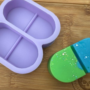 Può includere: Uno stampo in silicone viola a forma di lettera B con due cavità ovali. Un oggetto ovale bicolore verde e blu è appoggiato su una superficie di legno.