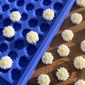 May include: A blue silicone mold filled with daisy-shaped resin pieces. The daisies are white with yellow centers. Some daisies are scattered on a wooden surface next to the mold. The image is well-lit, showcasing the details of the floral design.