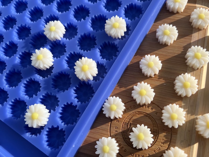 May include: A blue silicone mold filled with daisy-shaped resin pieces. The daisies are white with yellow centers. Some daisies are scattered on a wooden surface next to the mold. The image is well-lit, showcasing the details of the floral design.