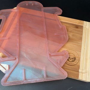 Può includere: Uno stampo in silicone rosa chiaro a forma di supporto per telefono, posto su un vassoio di legno con un logo a forma di teschio e il testo "Live With A Trail".