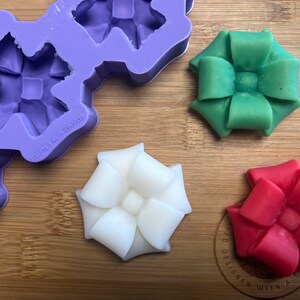 May include: Three handmade wax bow ornaments in green, white, and red. The ornaments are sitting on a wooden surface next to a purple silicone mould.