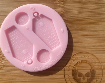 May include: Pink silicone mold with two coffin-shaped cavities, each featuring a crescent moon, stars, and an eye design. The mold also includes two round cavities, one with the word "YES" and the other with the word "ON". The mold is labeled "DESIGNED WITH A TWIST".
