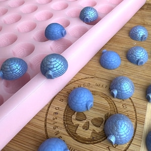 Könnte beinhalten: Eine Silikonform mit mehreren runden Vertiefungen, gefüllt mit blauen, glitzernden, igluförmigen Süßigkeiten. Die Süßigkeiten sind auch auf einer Holzoberfläche verstreut, mit dem Text "Designed with Love" sichtbar.