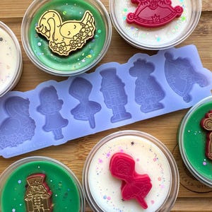 May include: A set of six wax melts in clear containers with various Christmas-themed designs. The melts are arranged on a wooden surface with a silicone mould for making wax melts. The designs include a swan, a nutcracker, a ballerina, a rocking horse, a mouse, and a Christmas tree.