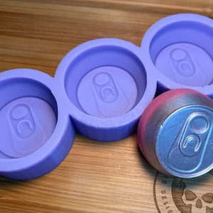 May include: A lilac-colored silicone mold with three circular cavities, each shaped like the top of a soda can. A finished, metallic-looking soda can is next to the mold. The mold has the text "DESIGNED WITH A BITE" on it. The items are on a wooden surface.