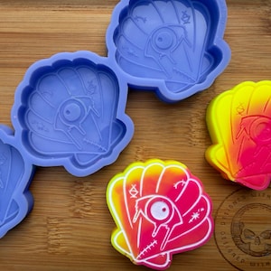 Puede incluir: Moldes de silicona azules con forma de concha y piezas de resina acabadas con forma de concha en amarillo, rosa y naranja. El diseño de la concha incluye un ojo, puntadas y otros detalles. El logotipo es una calavera.