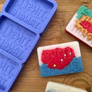 May include: A purple silicone mold with three rectangular cavities, each with the words "TINSEL TANGLE" embossed in the mold. Three finished soap bars are shown, each with the words "TINSEL TANGLE" in different colors and styles.