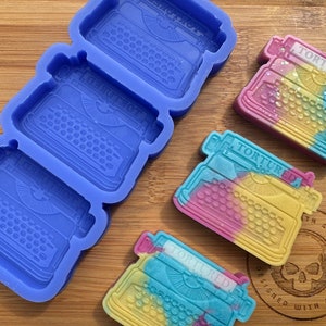 May include: Blue silicone molds and colorful typewriter-shaped soaps. The soaps are pastel-colored with the word "TORTURED" on the top. The soaps are yellow, pink, and blue. The molds are blue.