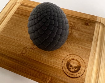 May include: A black dragon egg-shaped object with a textured surface, sitting on a wooden chopping board. The chopping board has a circular design with the text "Designed with a twist" and a skull graphic.