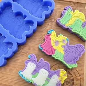 May include: A silicone mold for making ghost-shaped soaps or candies. The mold has three cavities, each shaped like a ghost with a hat and a broom. The mold is blue and has a textured surface. There are three finished ghost-shaped soaps in the image, each with a different color scheme.
